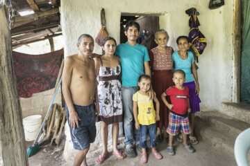 Auch Doña Glória (rechts hinten, 52 Jahre) und ihre Familie sind von der Krise betroffen- hier mit einem Teil ihrer Familie vor ihrem  Haus. Vor 40 Jahren haben sie aus der Not heraus Land besetzt. Bis heute können sie daher überleben. Viel ist es nicht, sie leben in extremer Armut. Neben dem fehlenden Land ist die nicht vorhandene Gesundheitsversorgung und Wasser ein Problem: es gibt immer weniger. Was ihr hilft ist ihr fester Glaube. Und Radio Progreso, das sie jeden Morgen hört – auch wenn der örtliche Priester dies verboten hat. Familienfoto v. links: Ehemann Pedro Muñoz, Sohn Omar Muñoz Lara ( 3.v.links) und  seine Frau Mary Selena Muñoz, Schwiegermutter Felipa Martinez, Gloria und 2 Enkelkinder